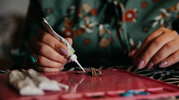 Close-up of hands applying glue to a small traditional craft piece on a pink surface, wearing floral attire
