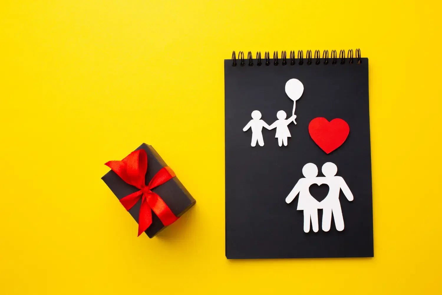 Black notebook with paper cutouts of family and red heart, next to black gift box with red ribbon on yellow background
