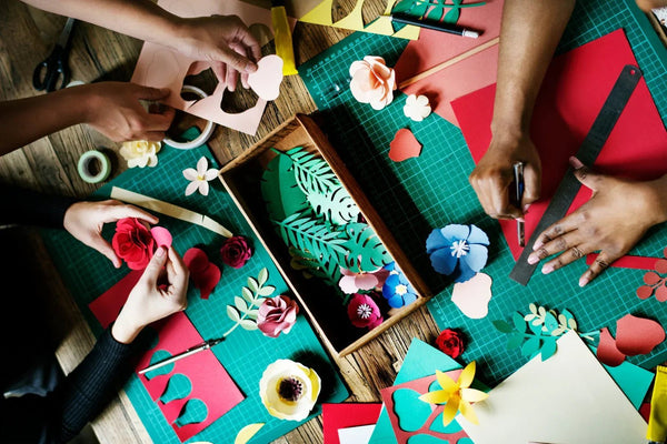 Hands crafting colorful paper flowers and leaves on green cutting mats with scissors and rulers