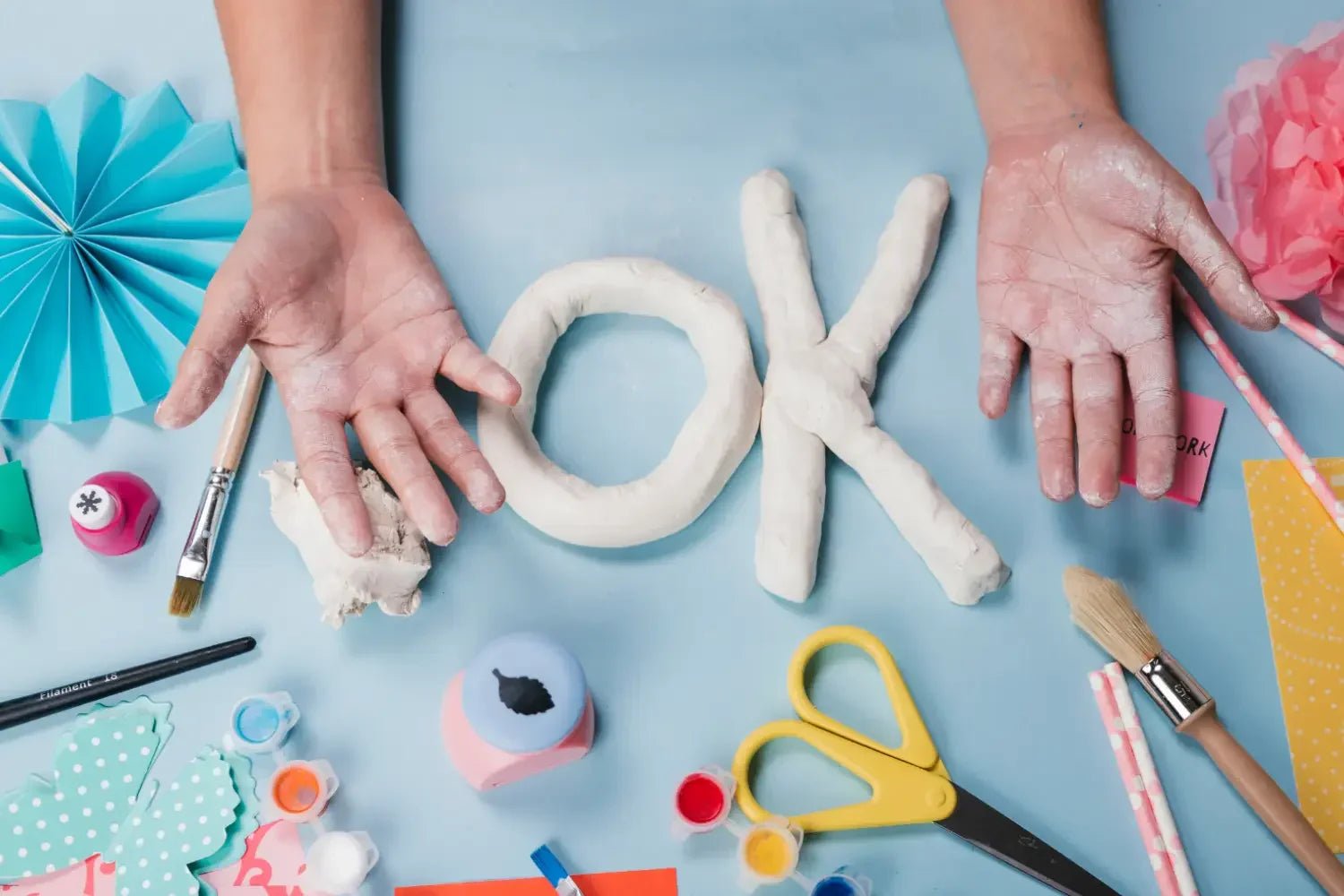 Hands covered in clay forming OK with white clay, surrounded by colorful craft supplies and brushes