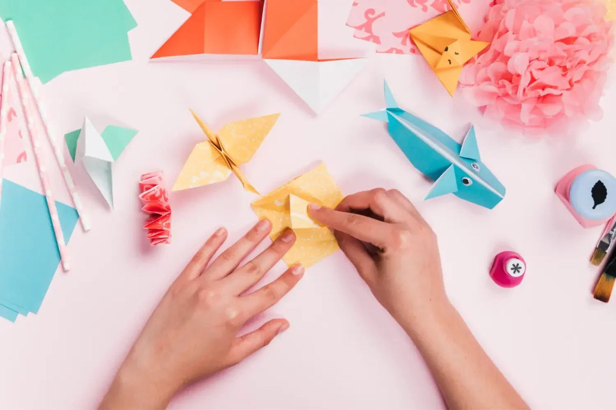 Hands folding yellow origami paper surrounded by completed origami animals and crafting tools