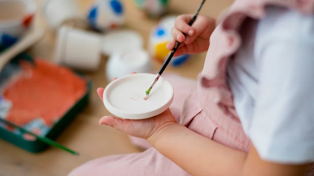 Close-up of child painting a small white ceramic plate with green and red paint, eco-friendly craft activity