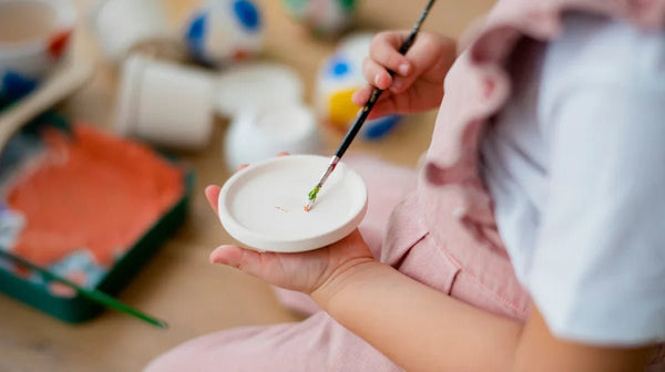 Close-up of child painting a small white ceramic plate with green and red paint, eco-friendly craft activity