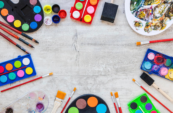 Top view of colorful watercolor palettes and paintbrushes arranged on a light wooden surface