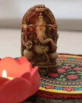 Intricately carved Lord Ganesha statue behind a lit red lotus tealight candle on colorful patterned mat